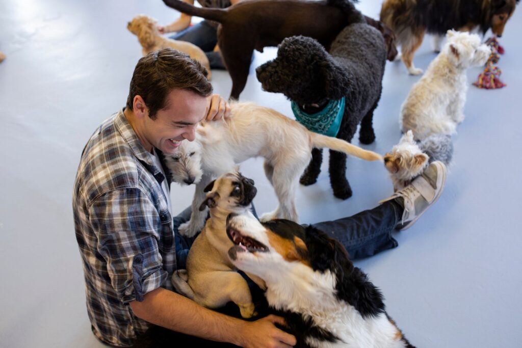 A smiling man sitting on the floor surrounded by a group of dogs